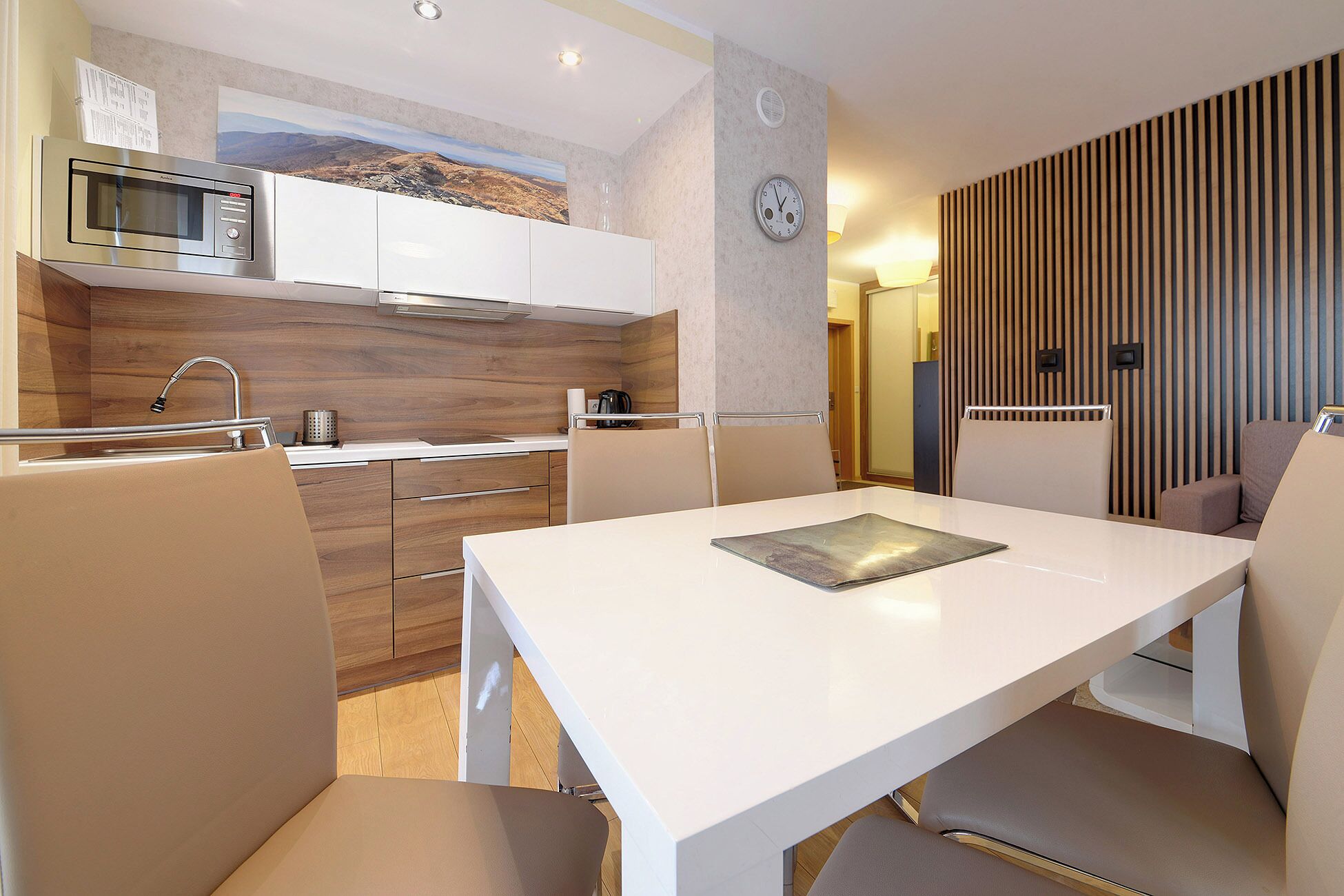 A modern kitchen with a microwave, stove, and cabinets. There is a dining table with chairs and a clock on the wall.