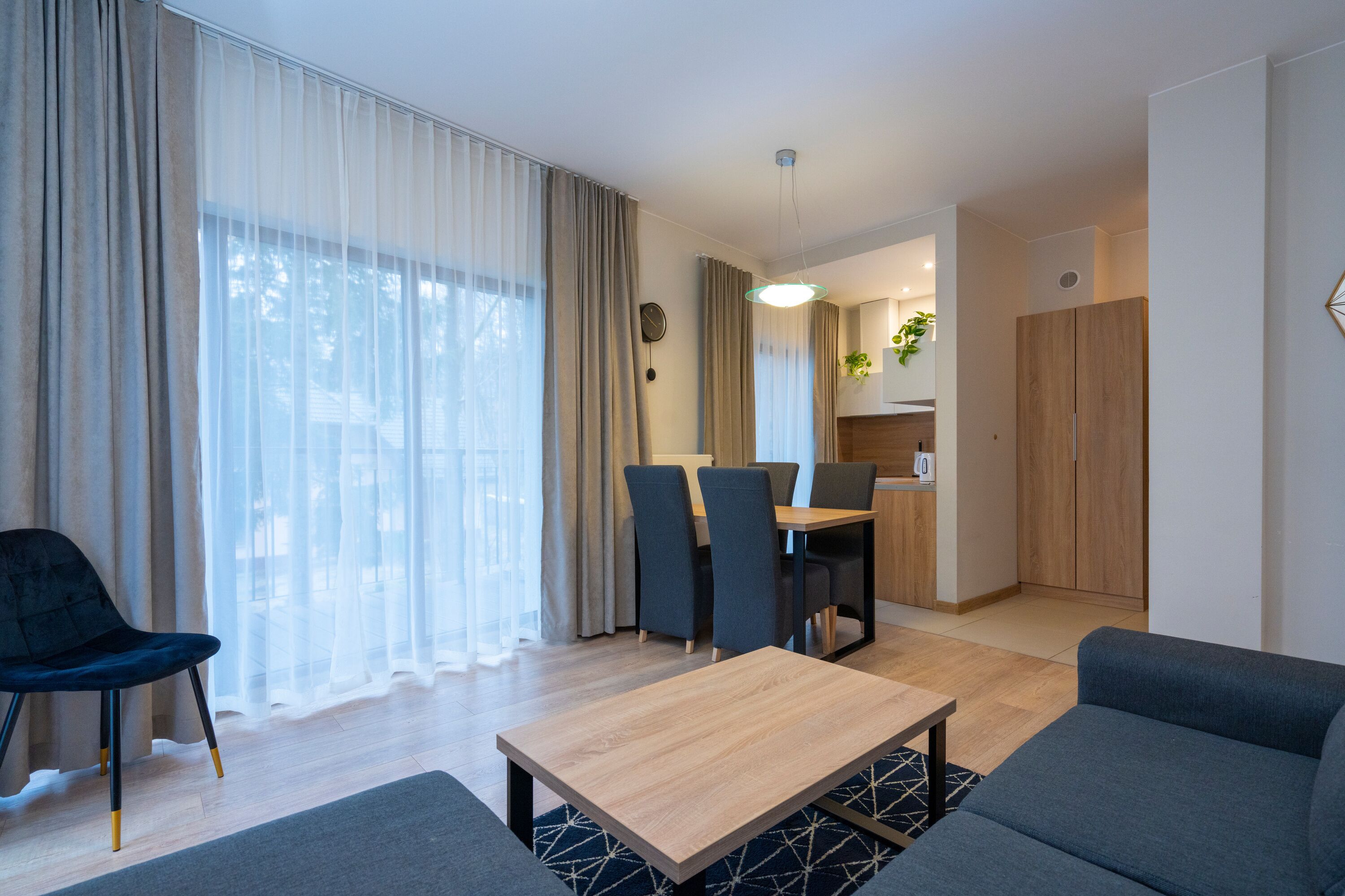Uma sala de estar moderna com um sofá, uma mesa de jantar e cadeiras. Ao fundo, há uma cozinha com um armário de madeira e uma planta.