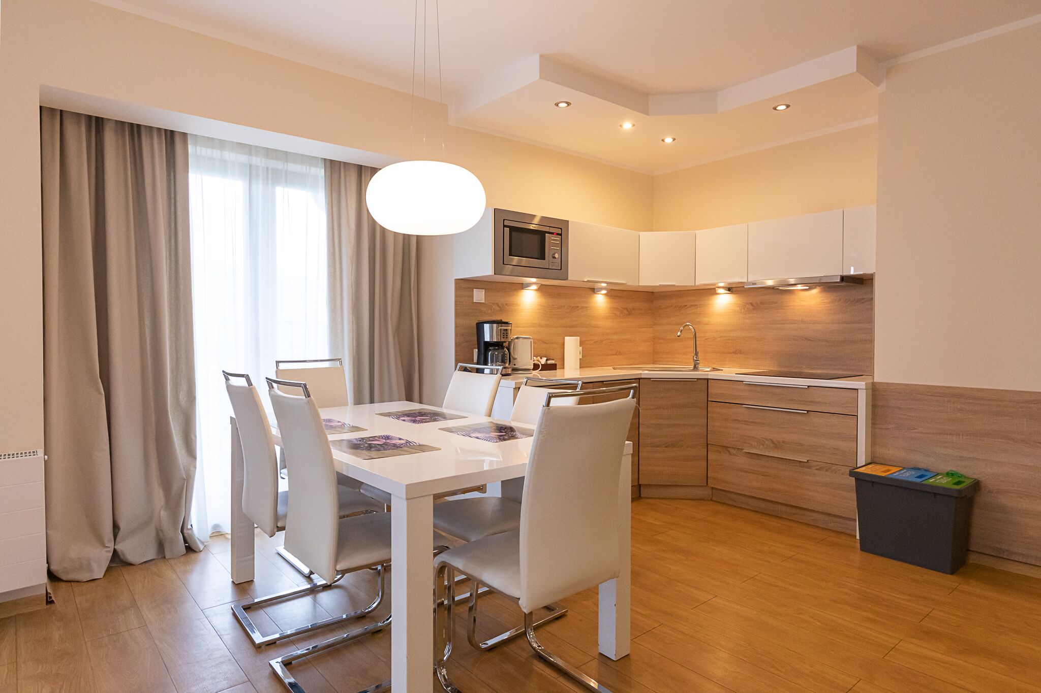 Una cocina moderna con armarios blancos, microondas y cafetera. Hay una mesa de comedor con sillas y un cubo de basura.