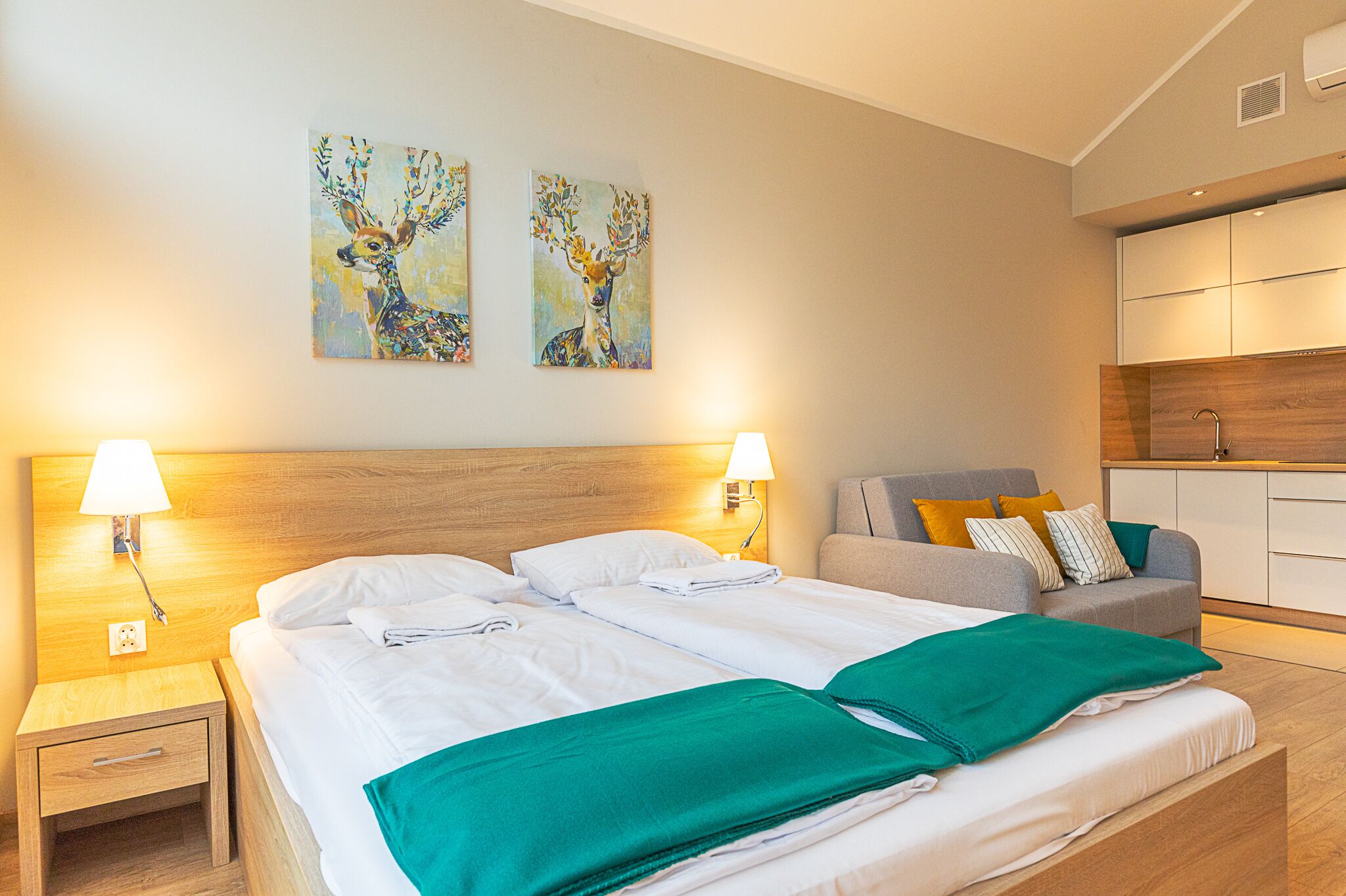 A hotel room with a large bed, two wall-mounted paintings, a bedside table with a lamp, a sofa with cushions, and a wooden headboard.