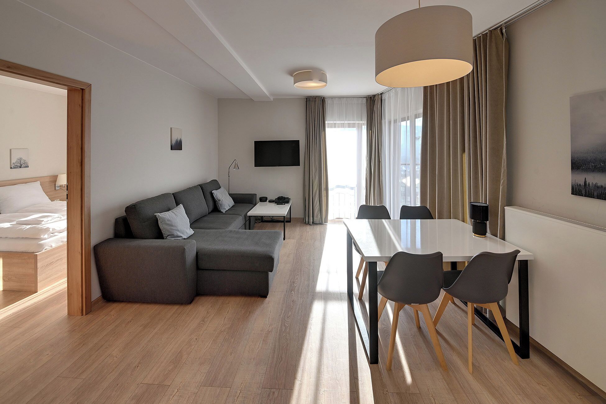 A modern hotel room with a wooden floor, a gray sectional sofa, a dining table with four chairs, a flat-screen TV mounted on the wall, and a large window with curtains.