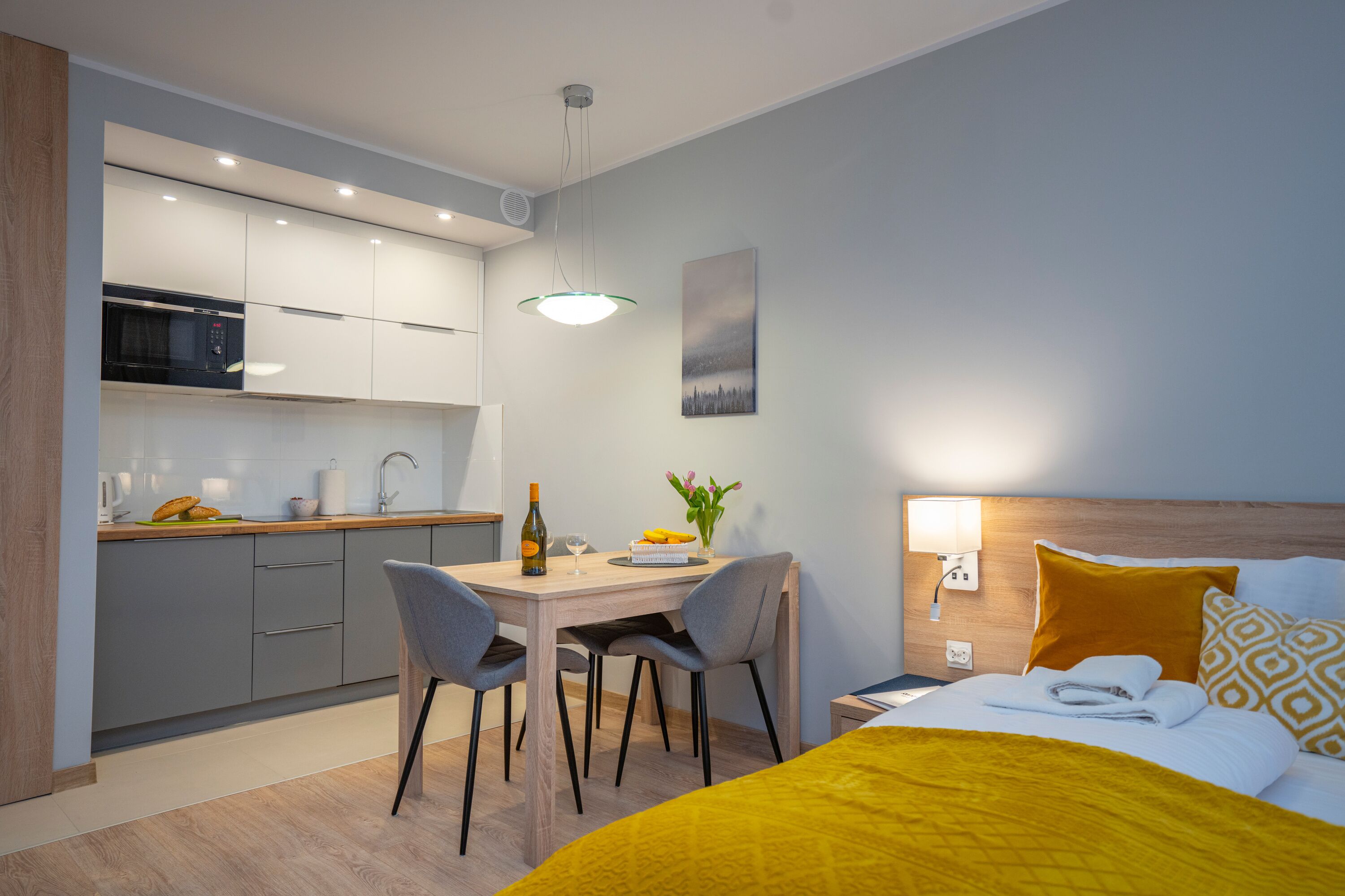 A modern hotel room with a bed, a bedside table, a nightstand, a lamp, and a view of the kitchen area.