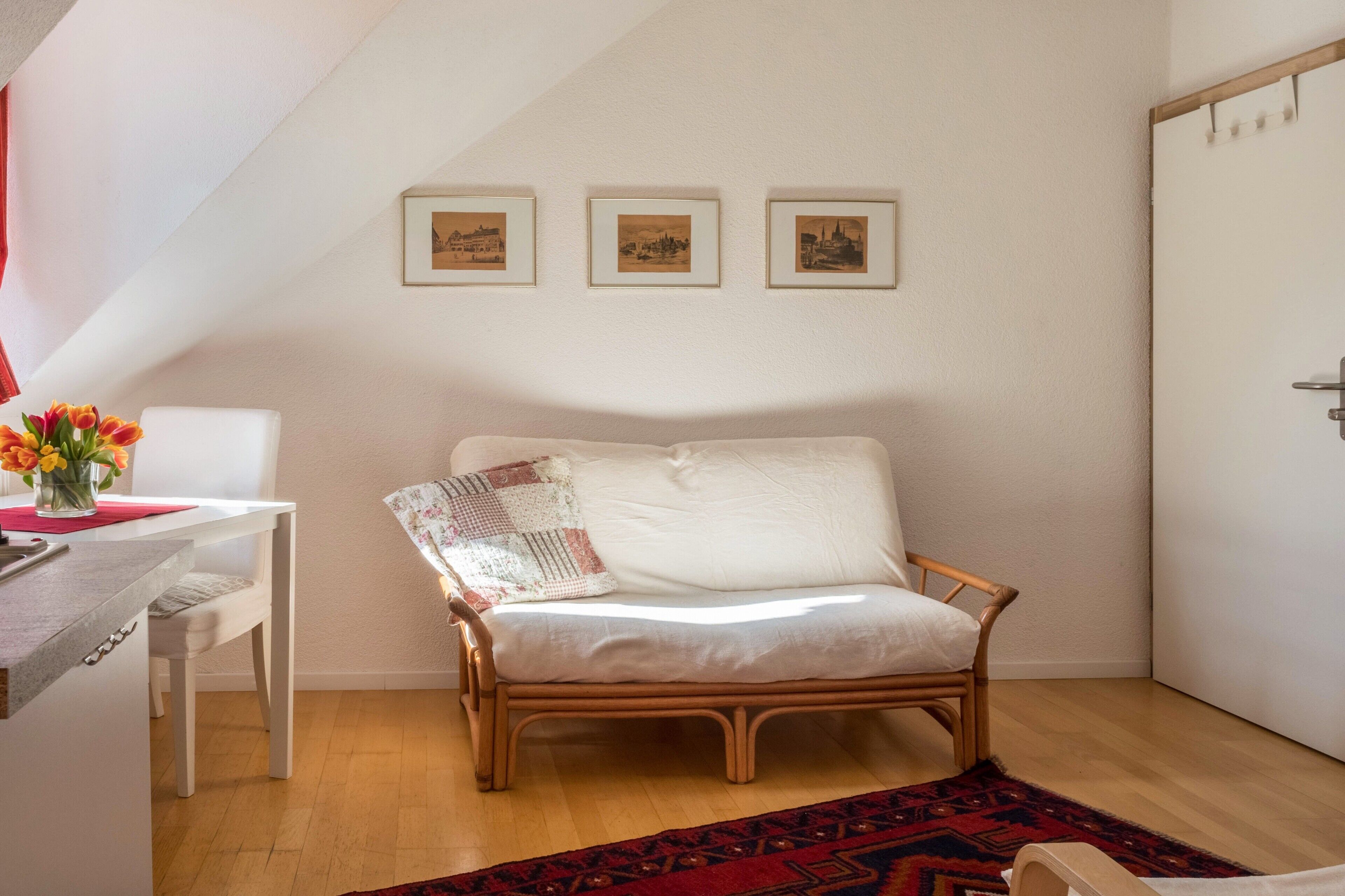 Una habitación acogedora con un sofá blanco, una mesita con un jarrón de flores y una silla de madera. Hay tres cuadros en la pared.