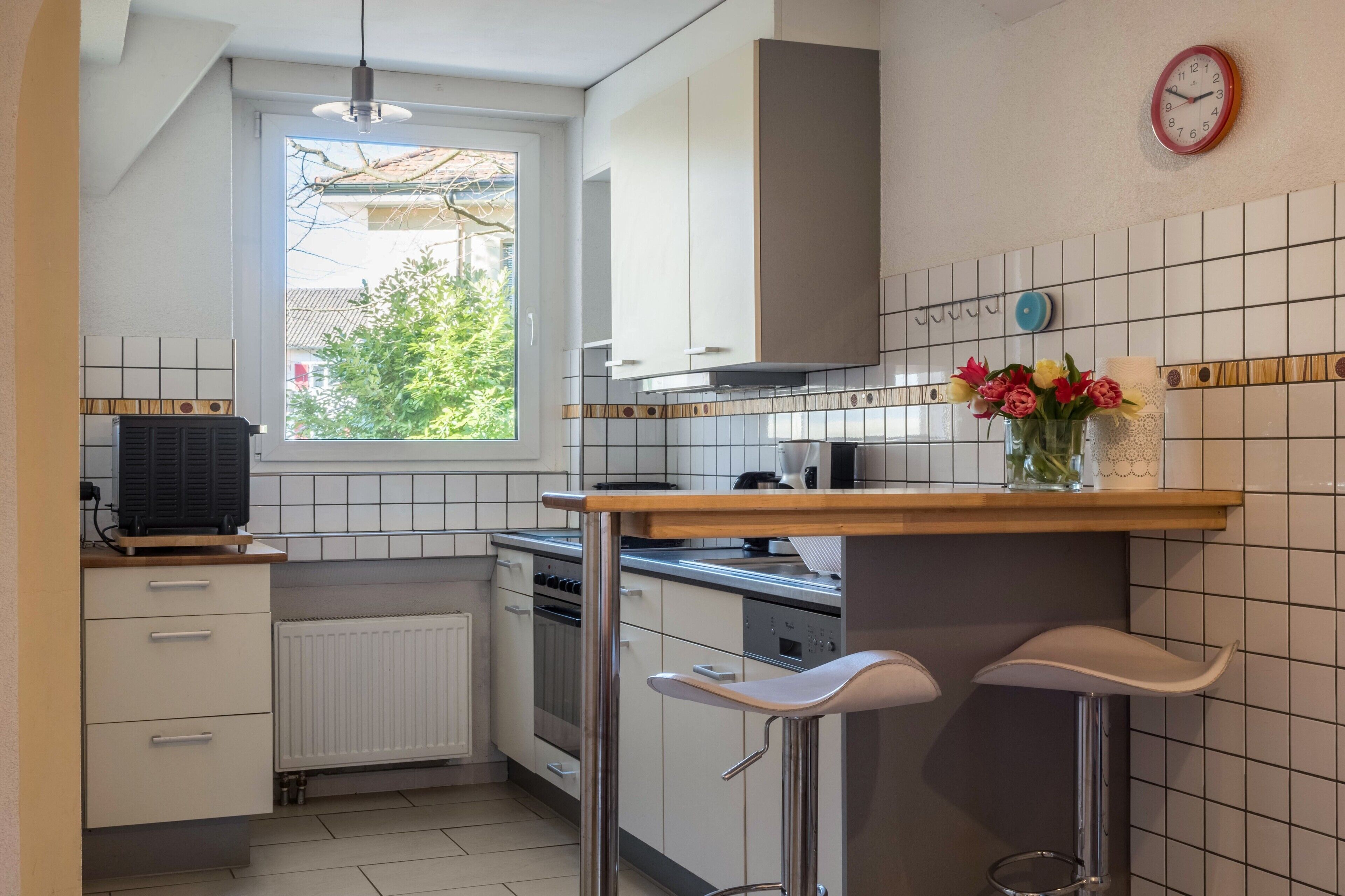 A kitchen with white cabinets, a window, a clock, and a bar area with stools.