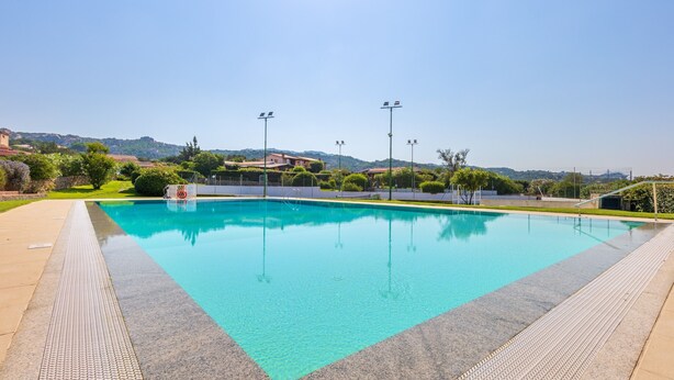 Una piscina rettangolare con superficie d'acqua cristallina e blu, circondata da un percorso pavimentato e un'area verde con alberi e arbusti.