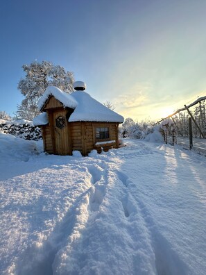 Lumihunnun peittämä puinen mökki, jonka katolla on lunta ja jossa on savupiippu. Mökkiä ympäröivät lumipeitteiset puut.