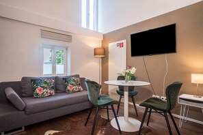Un salon moderne avec un canapé gris, des fauteuils verts, une table ronde ornée d’un vase, un téléviseur à écran plat et une fenêtre avec des stores.