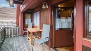 Een houten balkon met een tafel en stoelen, een raam met gordijnen en een deur naar binnen.