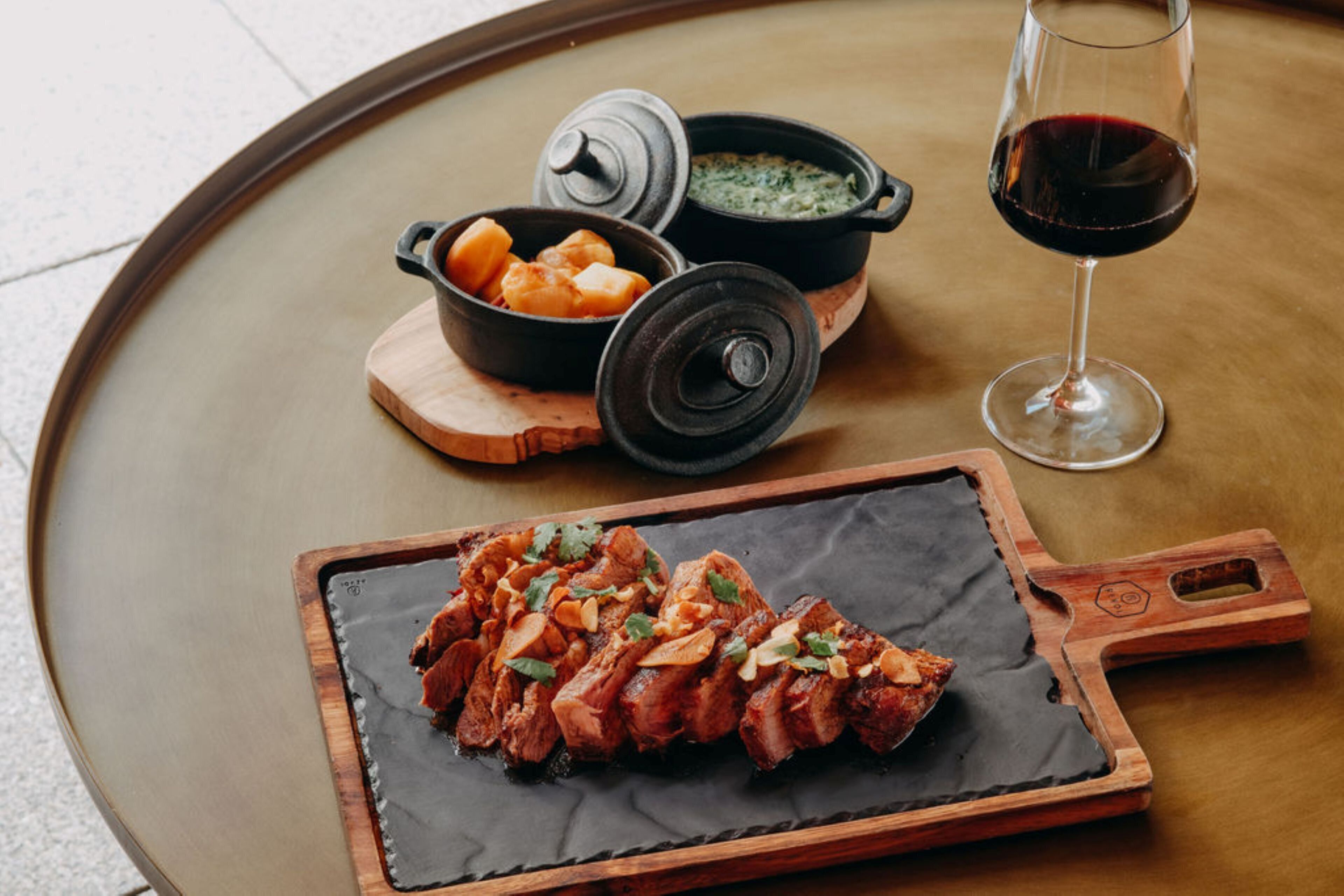 A round wooden table with a black slate serving board, a glass of red wine, and two black pots with lids.