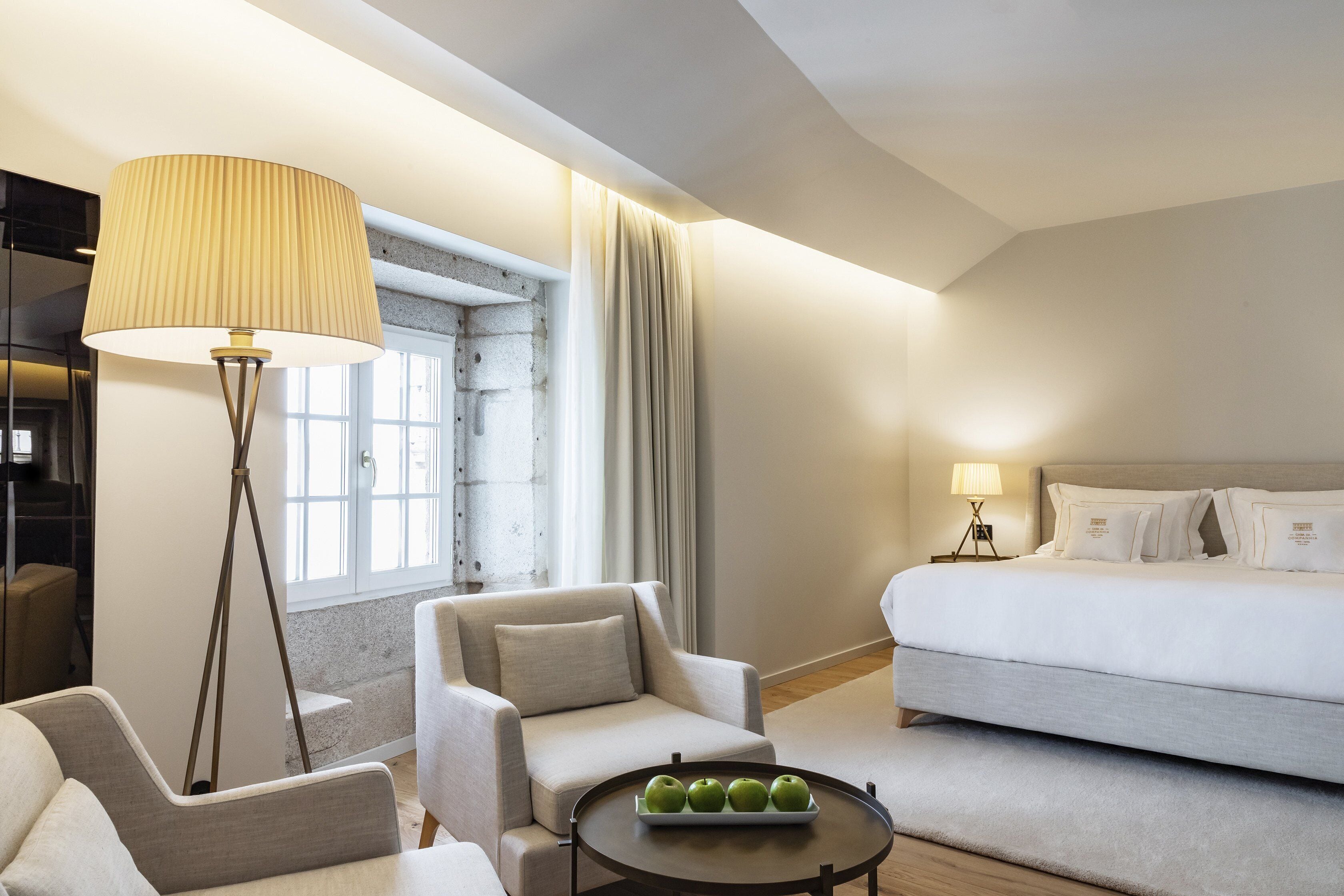 A modern hotel room with a sofa, a chair, a bedside table with a lamp, a bed, and a window with curtains.