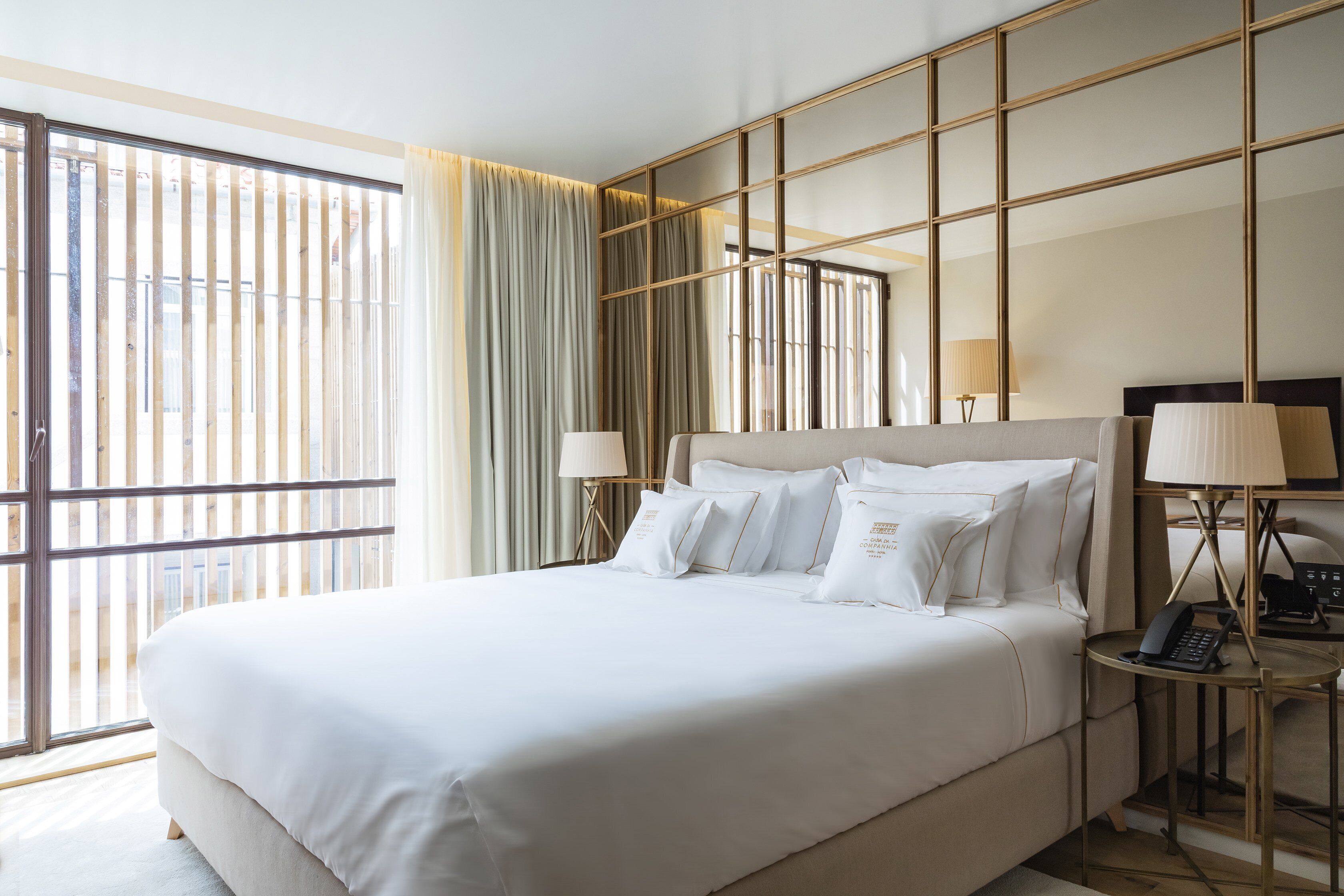 A hotel room with a large bed, two bedside tables with lamps, a large window with blinds, and a view of the outside.