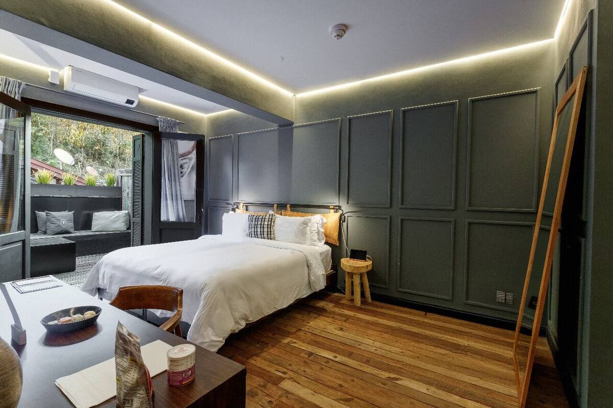 Une chambre moderne avec un grand lit, un plancher en bois, une petite table avec un bol et un sac, et une vue sur la verdure à l’extérieur.