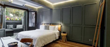 A modern bedroom with a large bed, a wooden floor, a small table with a bowl and a bag, and a view of greenery outside.
