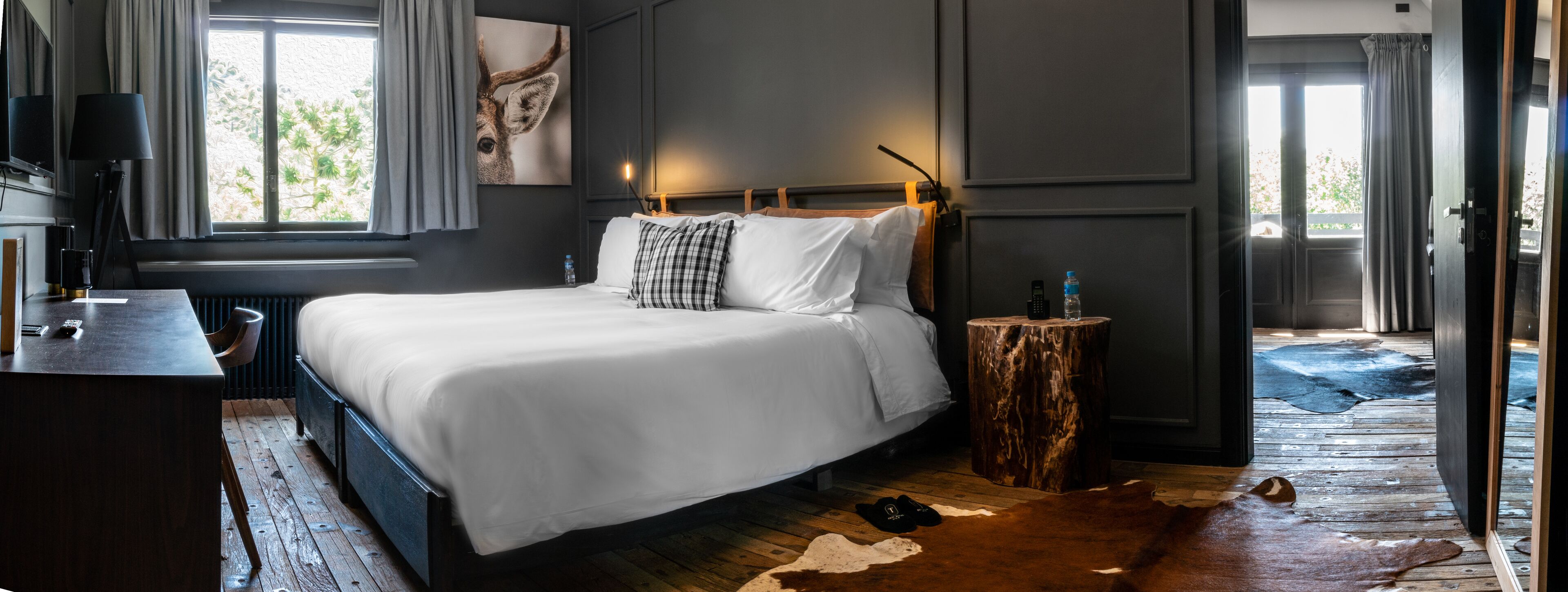 Un dormitorio moderno con una cama grande, una mesita de noche de madera, un escritorio con lámpara y vistas al exterior a través de una ventana.