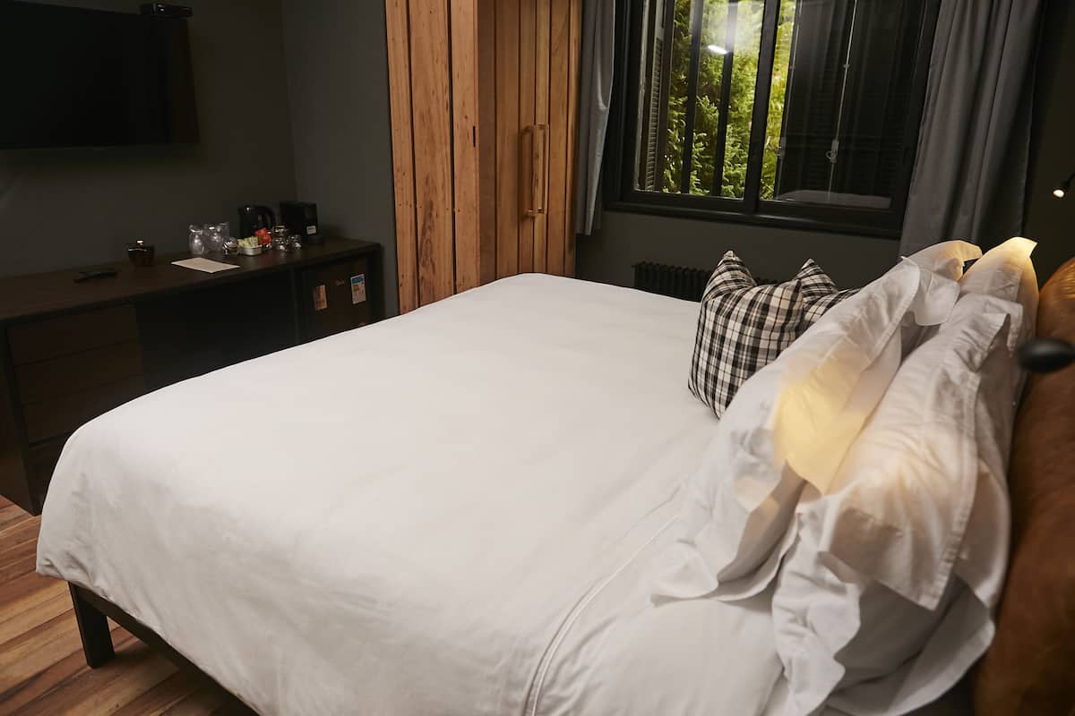 Un lit bien fait, avec du linge de lit blanc, une table de chevet en bois sur laquelle se trouve une machine à café, et une fenêtre avec des rideaux.