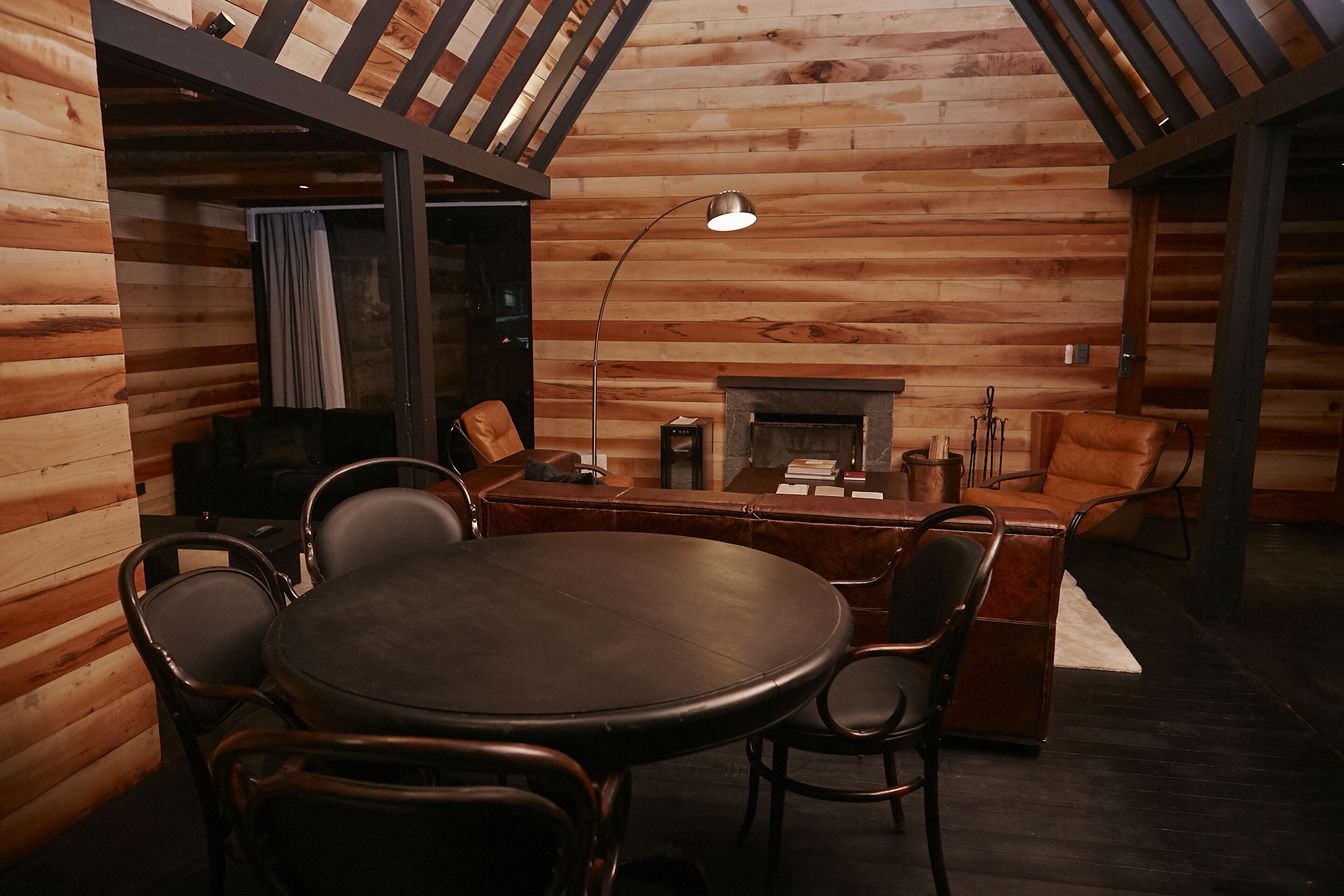 A cozy cabin interior with a round wooden table, leather chairs, a fireplace, and a large window with curtains.