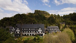 Un grand bâtiment de style traditionnel, doté de nombreux balcons, entouré d’une végétation luxuriante et bordé d’une colline boisée.