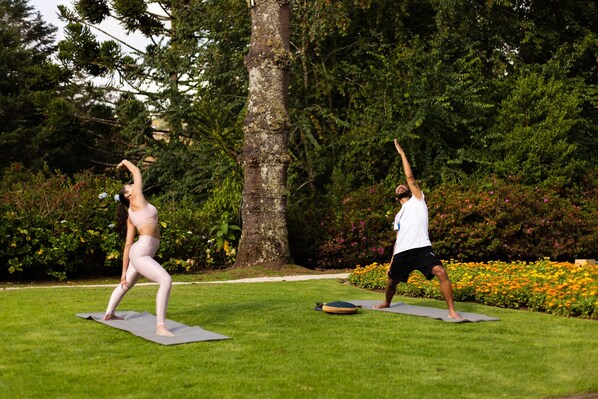 Two people practicing yoga on mats in a grassy outdoor area with trees and flowers in the background.