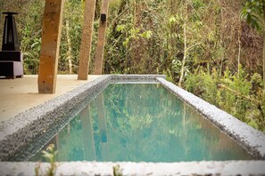 Une piscine rectangulaire dont les bords sont recouverts de petits galets, entourée d’une végétation luxuriante et de piliers en bois.