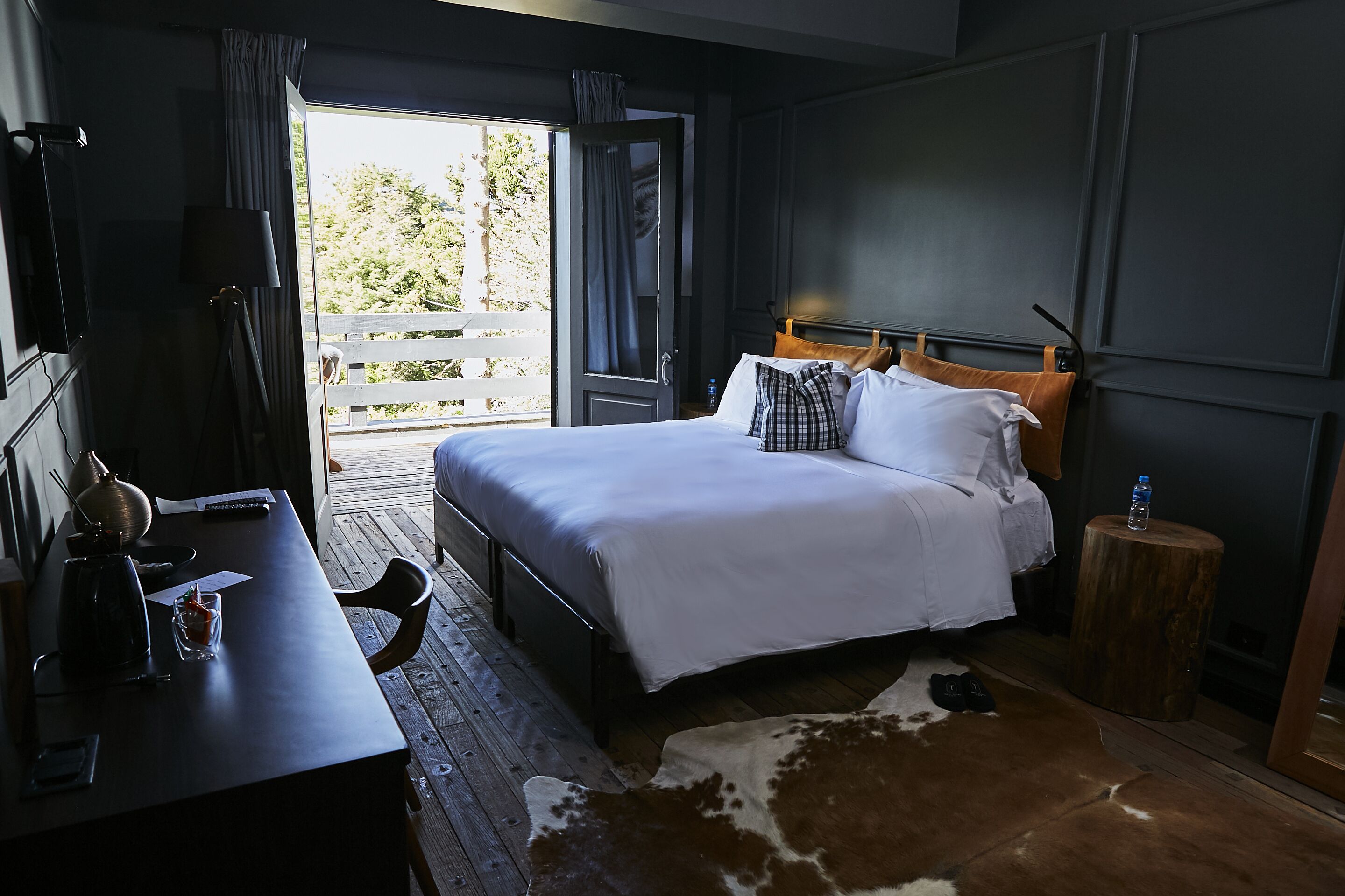 Habitación de hotel con cama, escritorio de madera, una mesita redonda, alfombra de cuero de vaca y balcón con vistas a la vegetación.
