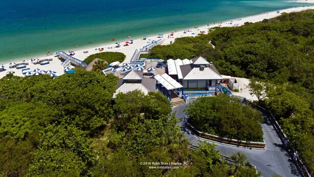 Vista aérea de um resort à beira-mar, com uma visão nítida do oceano, uma praia de areia e um estacionamento com vários veículos.