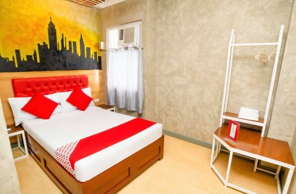 A hotel room with a bed, a red headboard, a cityscape mural, a window with curtains, and a white bedside table.