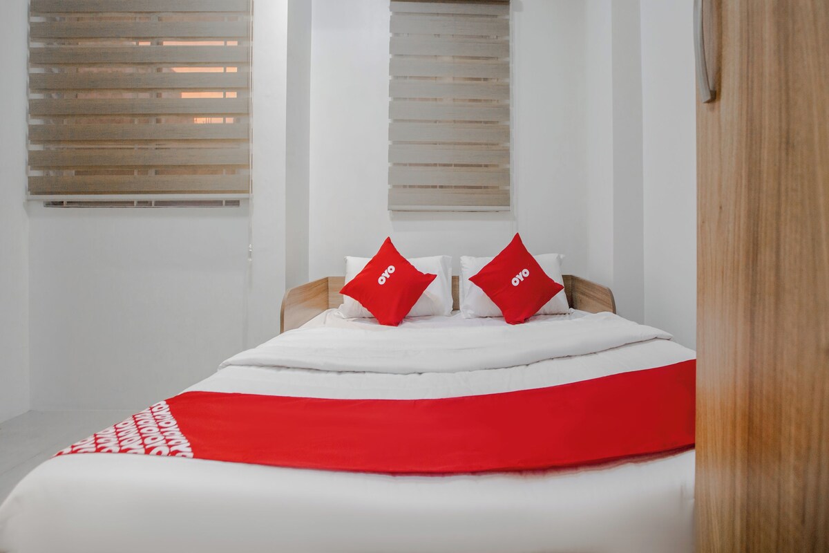 A bedroom with a bed, two windows with blinds, and a wooden door.