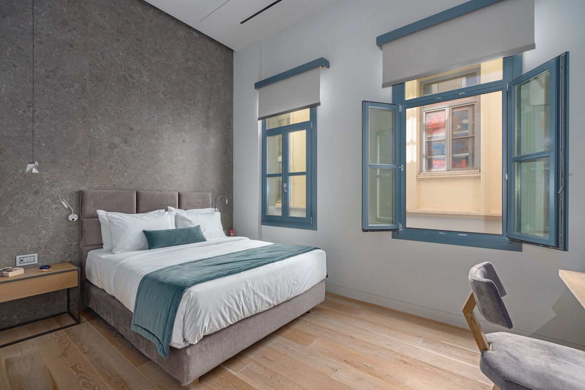 A modern bedroom with a bed, bedside table, chair, and large windows.