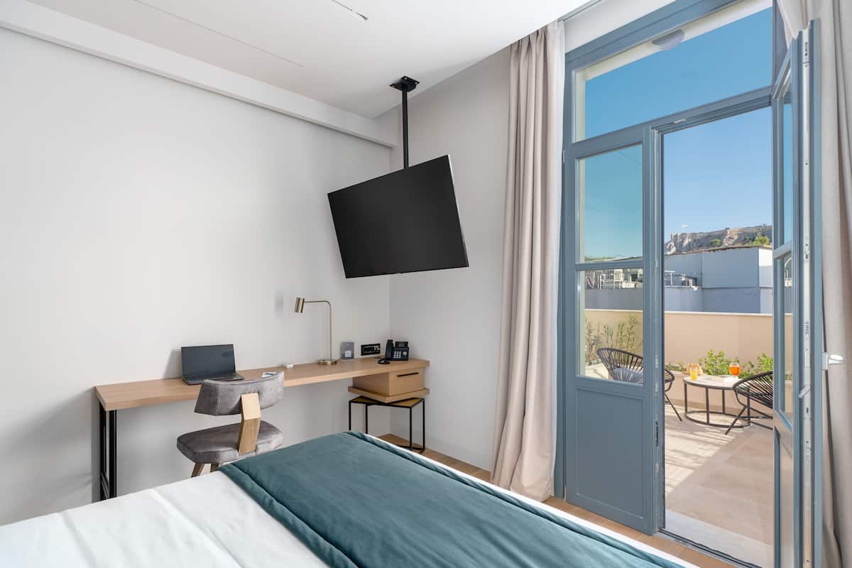 A modern hotel room with a bed, a desk with a laptop, a flat-screen TV, and a balcony with outdoor seating.