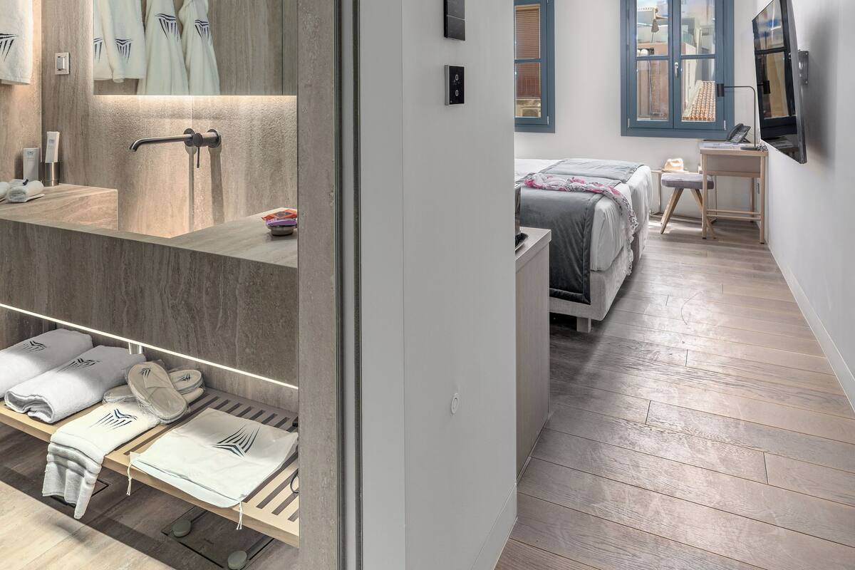 A modern hotel room with a large mirror, a wooden floor, a bed with a gray bedspread, a bedside table with a lamp, and a window with blue shutters.