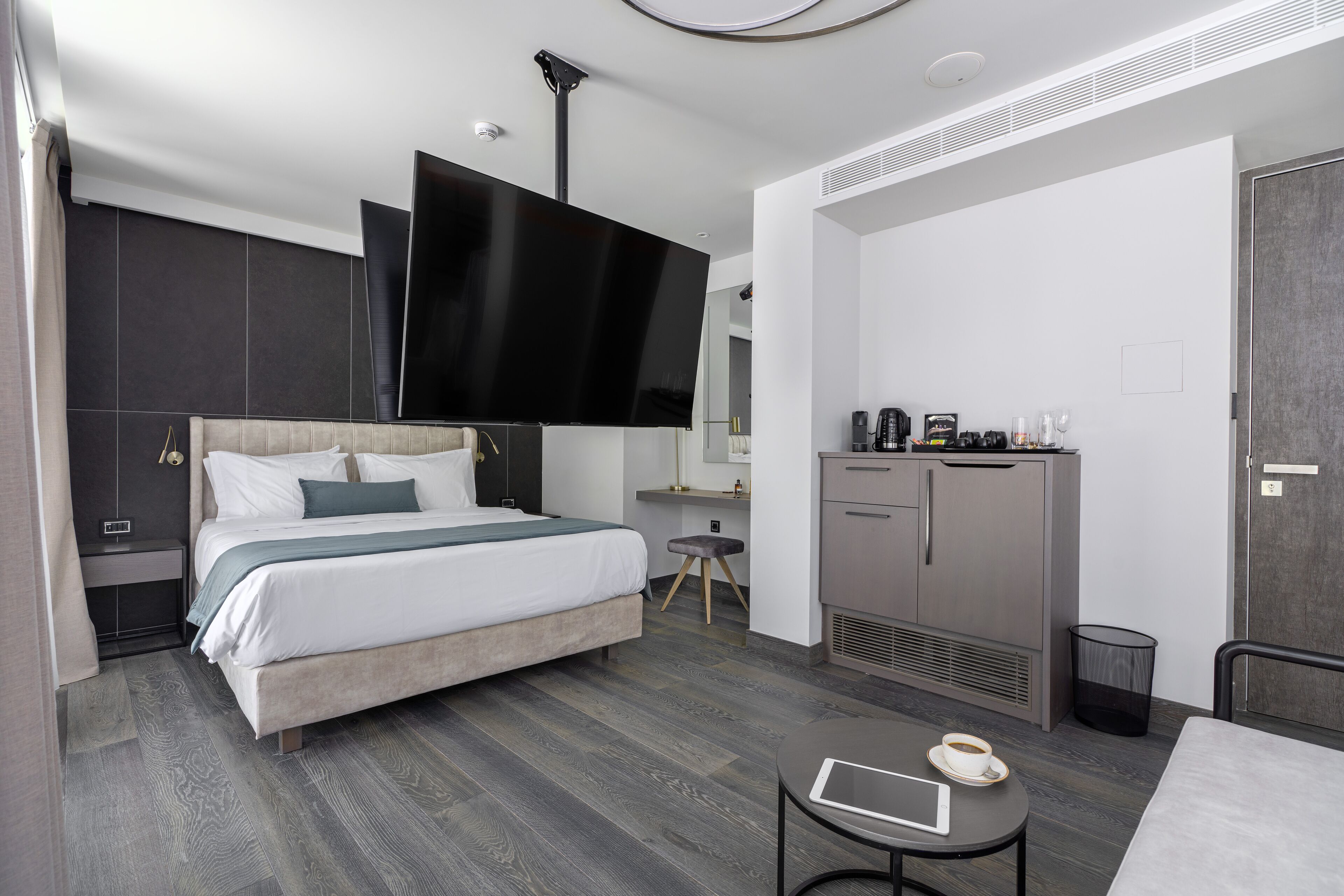 A modern hotel room with a large bed, a desk with a computer, a coffee machine, and a flat-screen TV mounted on the wall.