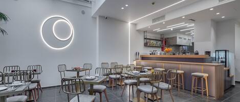 A modern restaurant with a central bar, multiple dining tables with chairs, and a circular light fixture on the wall.