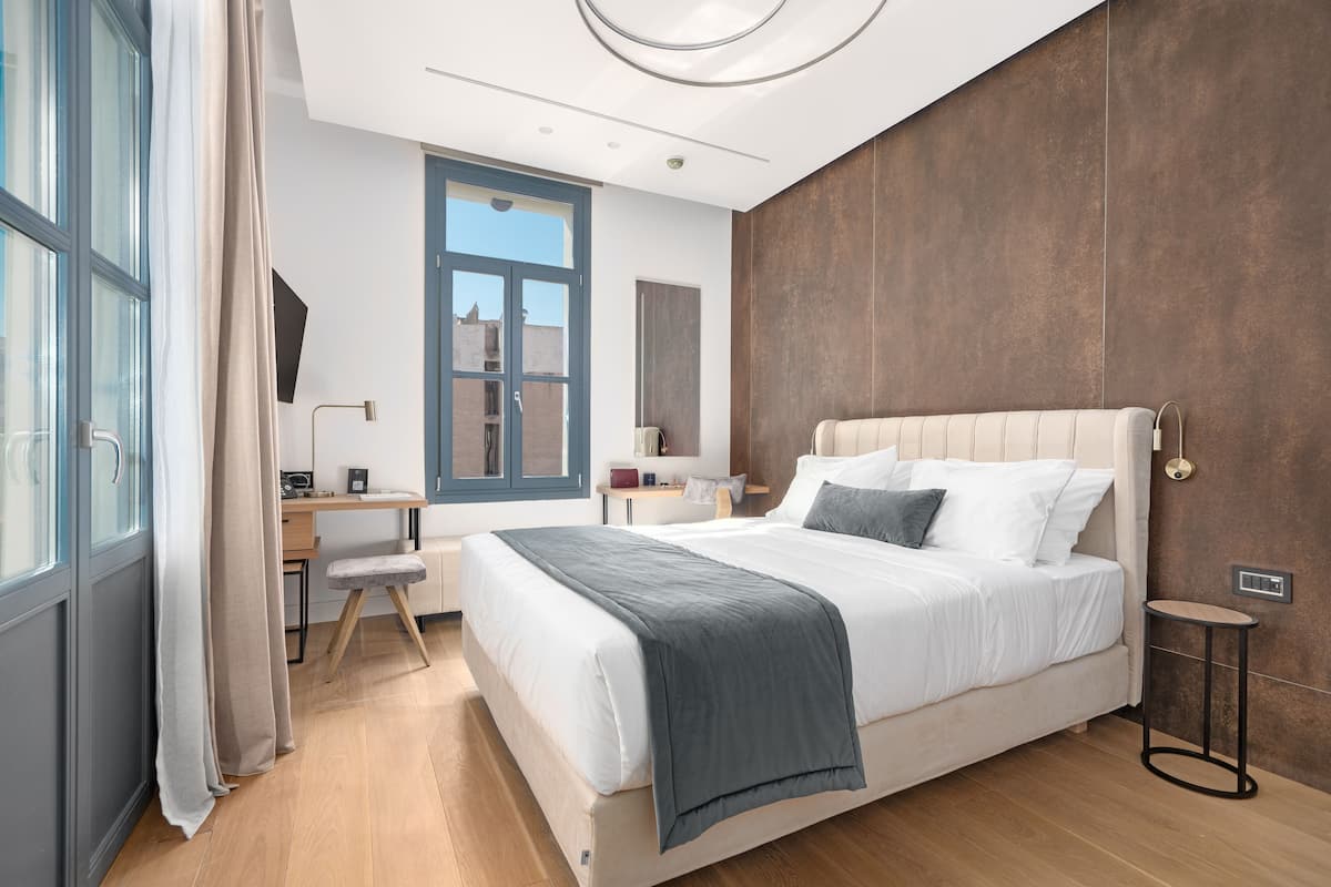 A modern bedroom with a large bed, a desk, and a view of the city through a window.