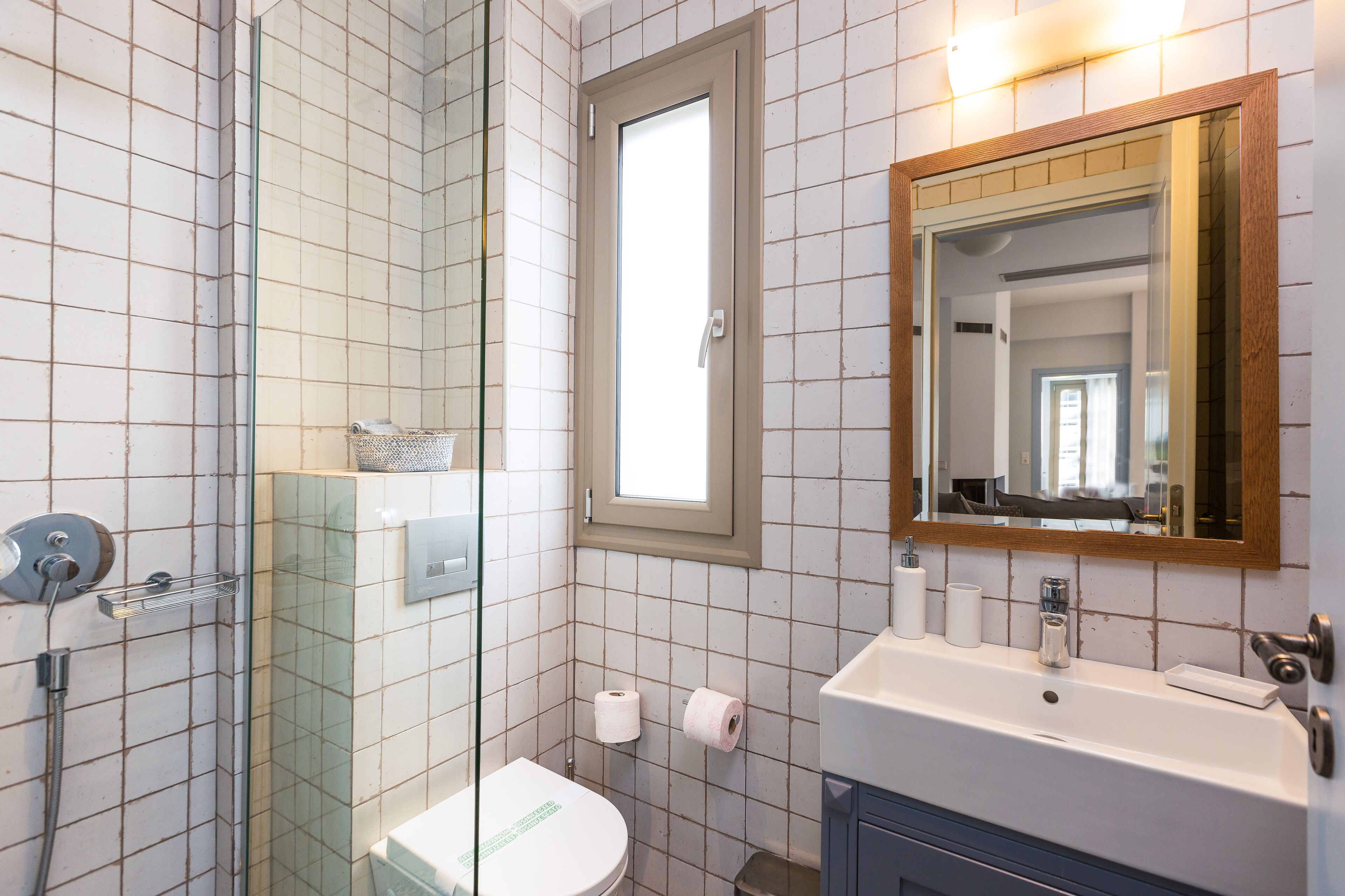 Une salle de bain avec une douche à parois de verre, des toilettes, un lavabo avec un miroir et une fenêtre située au-dessus des toilettes.