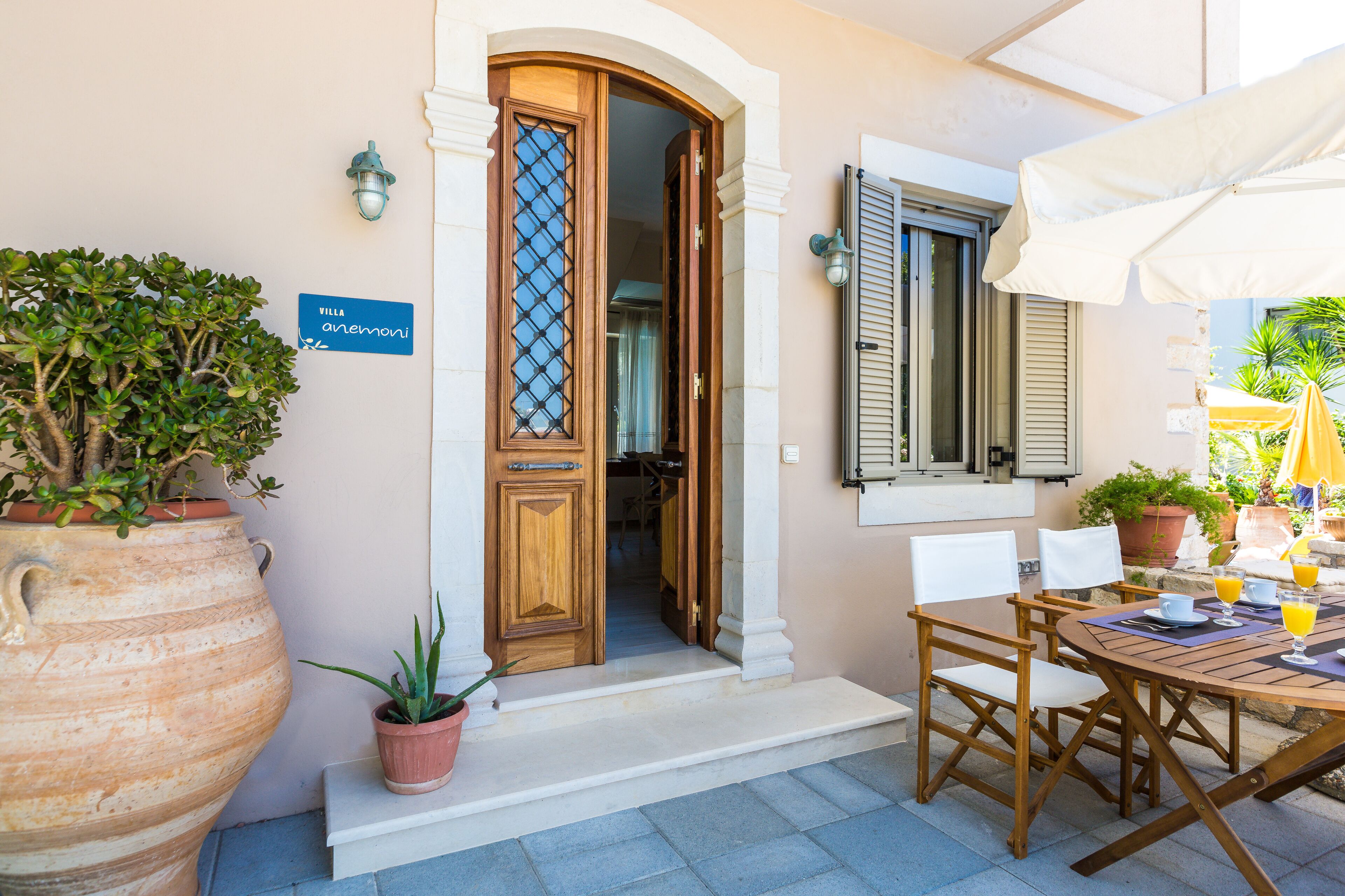 A villa entrance with a wooden door, a sign reading "Villa Anemoni," outdoor seating with a table and chairs, and potted plants.