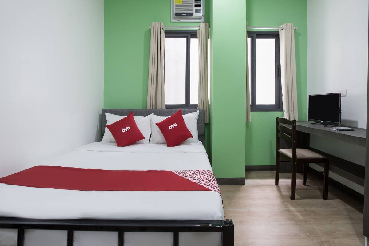 A hotel room with a bed, a desk, and two windows.