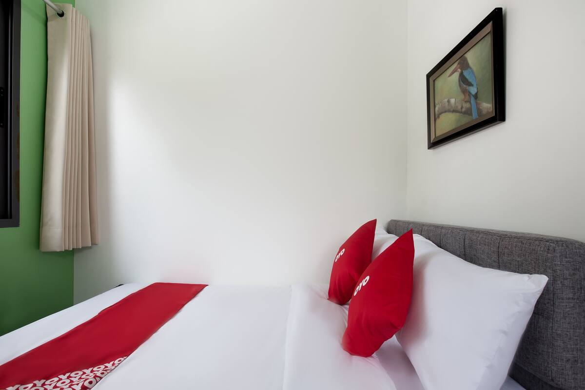 A hotel room with a bed, red and white pillows, a grey headboard, a painting on the wall, and a window with curtains.