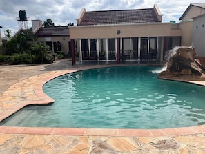 A swimming pool with a curved edge, a water fountain, and a covered seating area with chairs.