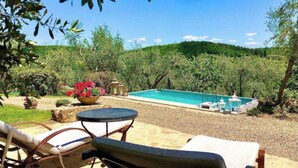 Un espace piscine doté de chaises longues et d’une table, entouré de verdure et de collines.