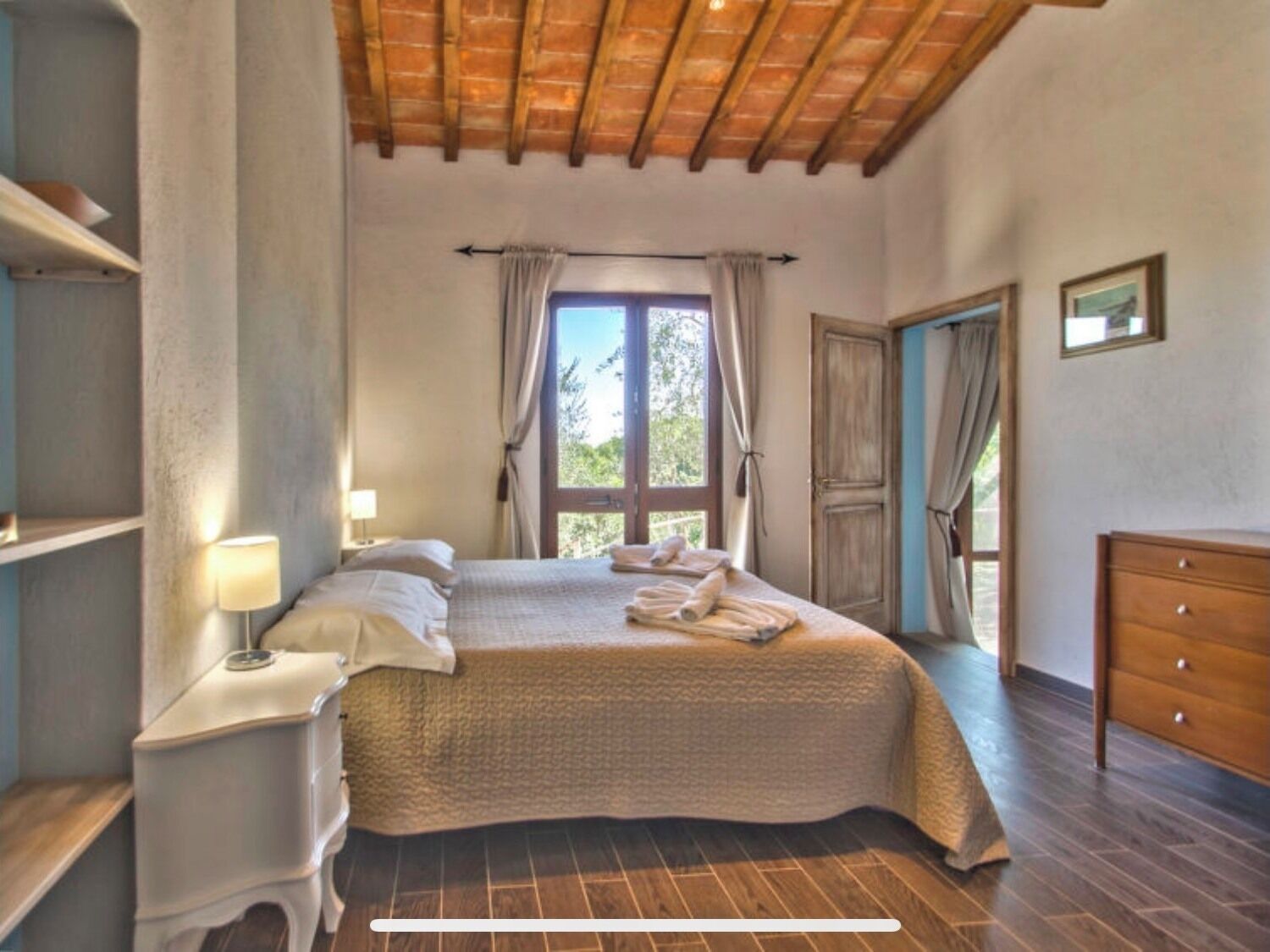 Un dormitorio con cama, mesita de noche con lámpara, cómoda, ventana con cortinas y puerta.