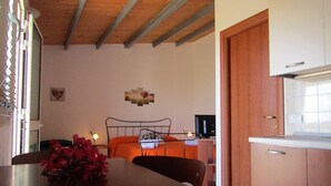 Un dormitorio con cama, mesita de noche y un escritorio pequeño con silla. Hay una ventana con persianas, un televisor montado en la pared y una puerta que da a una zona de cocina.
