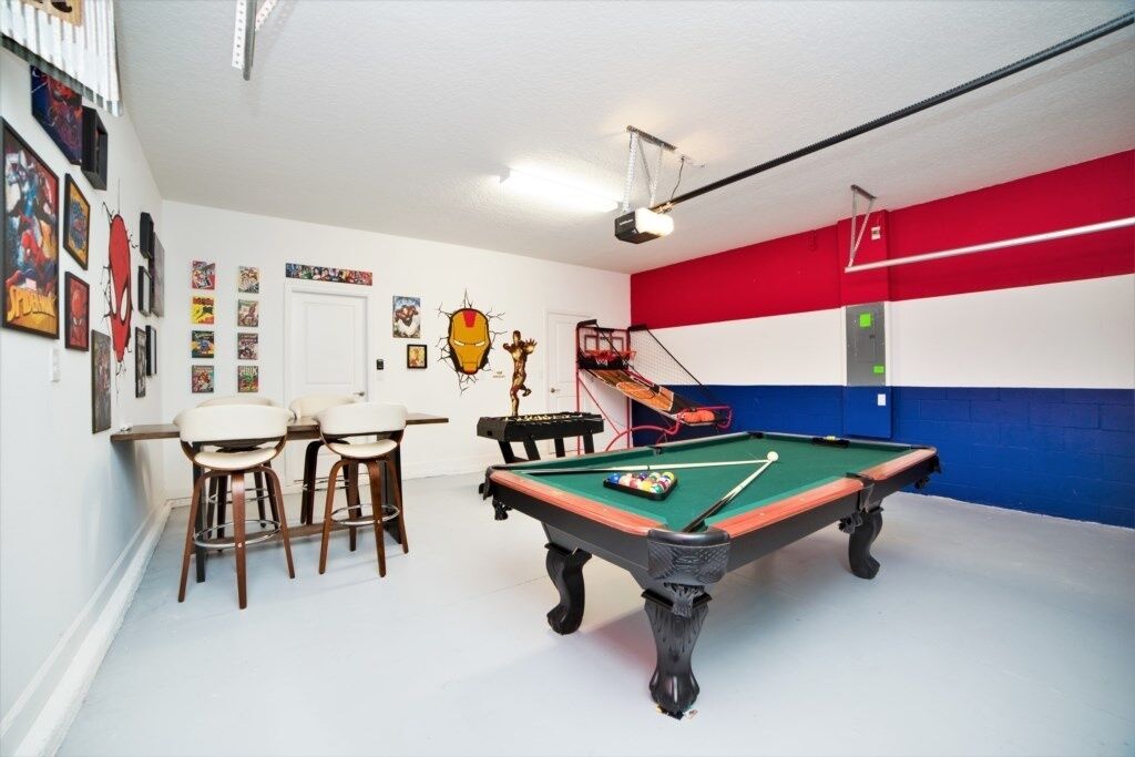 A pool table with a green felt surface, surrounded by white walls with various framed pictures. There is a red and blue wall panel, and a white door in the background.