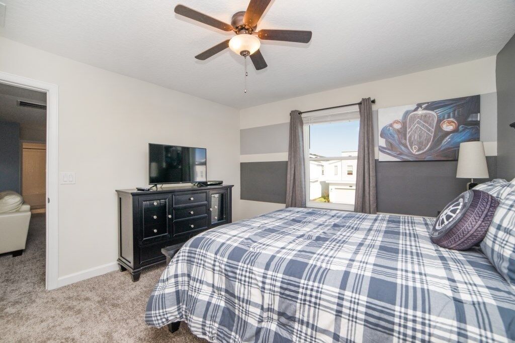A bedroom with a bed, a ceiling fan, a TV on a dresser, and a window with curtains.