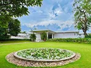 Um edifício moderno com um lago circular em primeiro plano, rodeado por um gramado bem cuidado e diversas plantas.