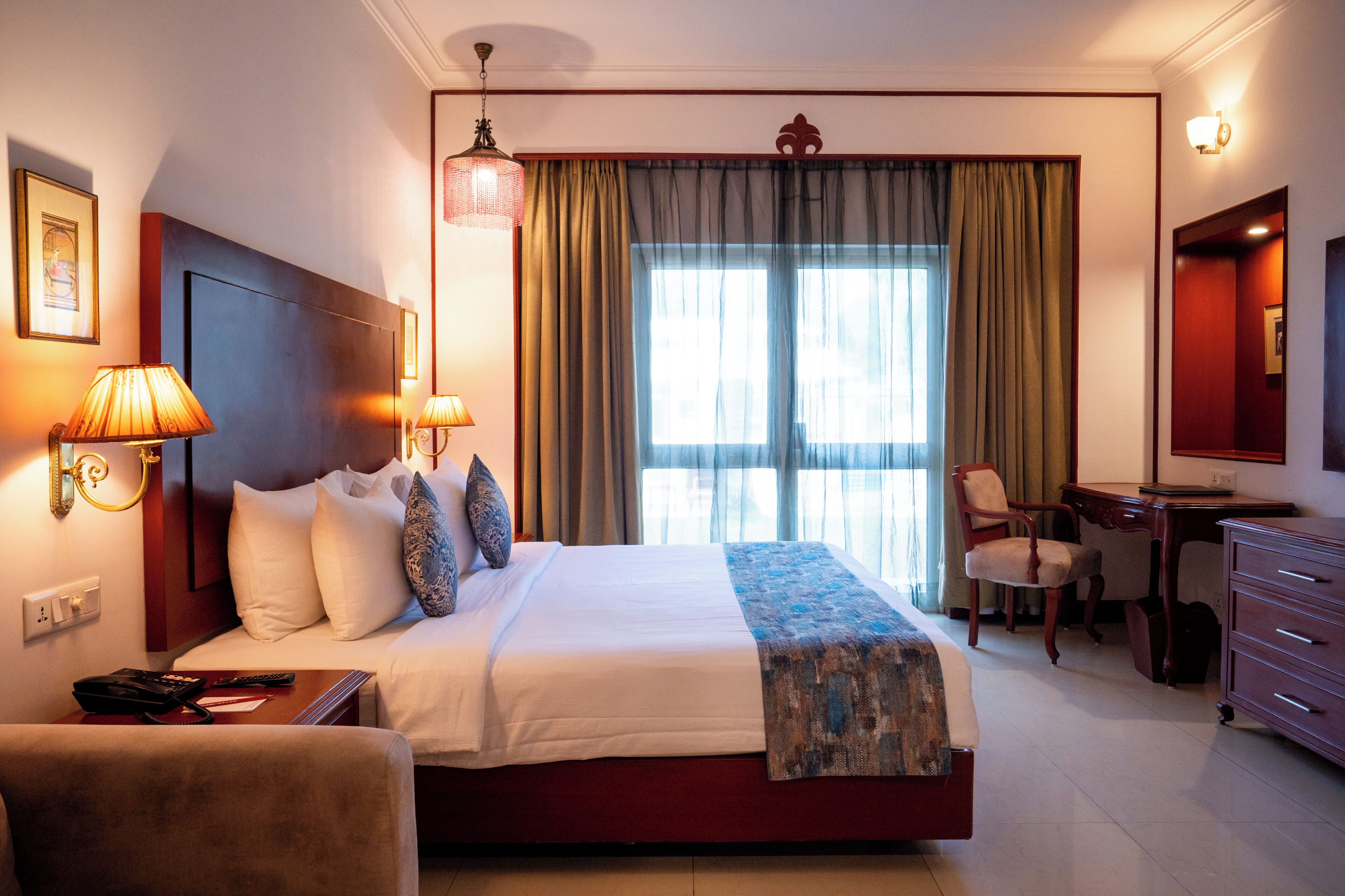 Une chambre d’hôtel comprenant un lit, des lampes de chevet, un bureau, une chaise, une commode et une grande fenêtre avec des rideaux.