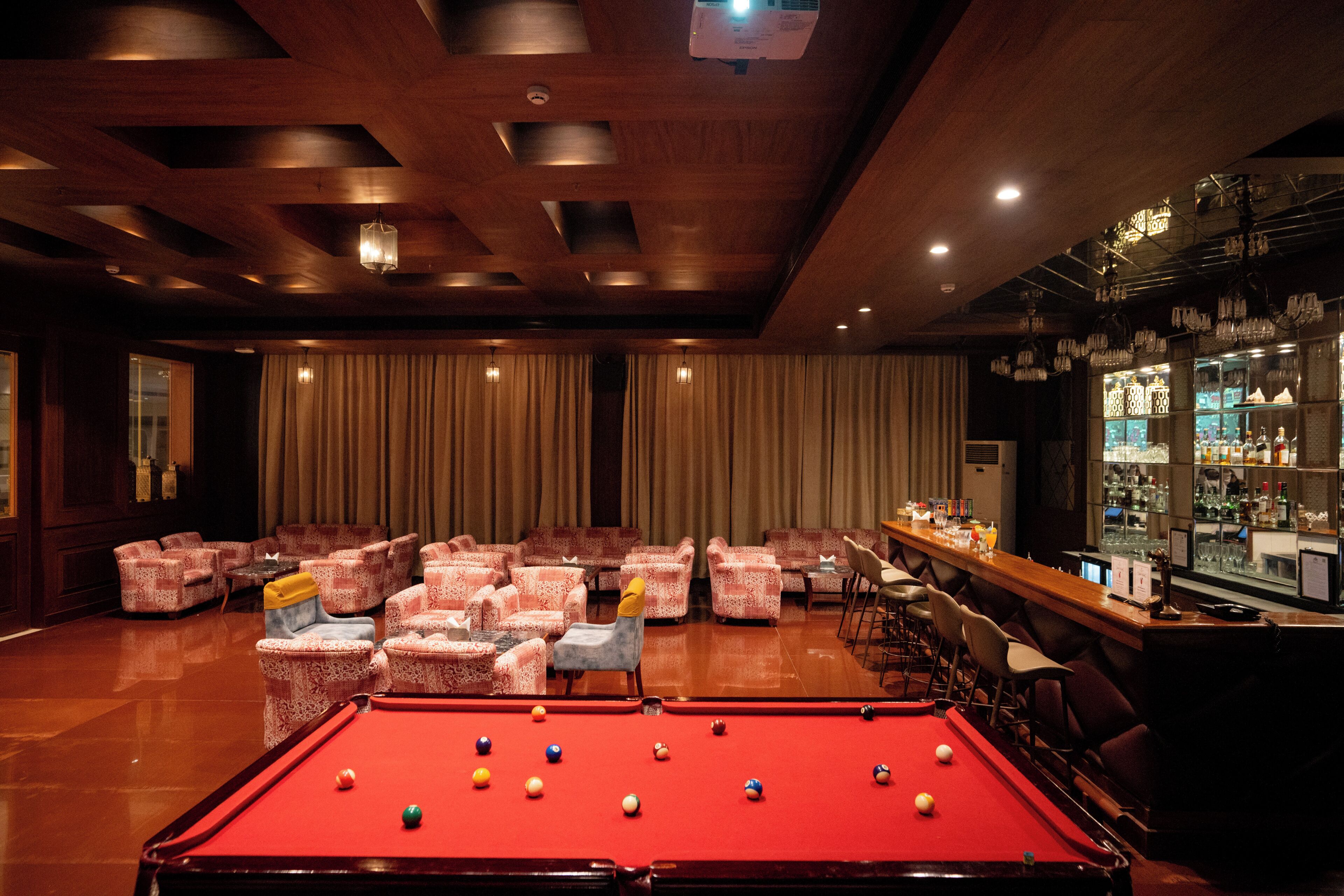 A pool table with balls, surrounded by patterned chairs, a bar area with bottles, and a bar counter.