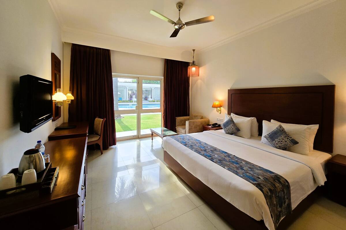 Habitación de hotel con cama, televisor, escritorio con cafetera, silla y vista a una piscina y zonas verdes.