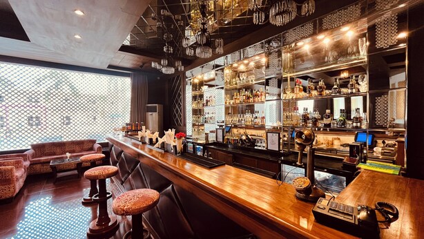 Un bar con una barra larga, varios taburetes y una variedad de botellas y vasos en estantes.