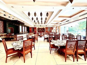 A spacious restaurant with multiple dining tables and chairs, a kitchen area, and a view of the outdoors. - Hotel Chandela Khajuraho (Rajnagar)