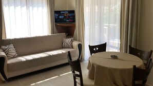 Uma sala de estar com sofá, cadeiras, uma mesa e uma televisão exibindo 'DRAMA'.